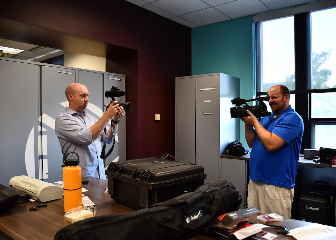 Two men hold cameras and face each other. There is a tall table in front of them with equipment cases and office supplies.